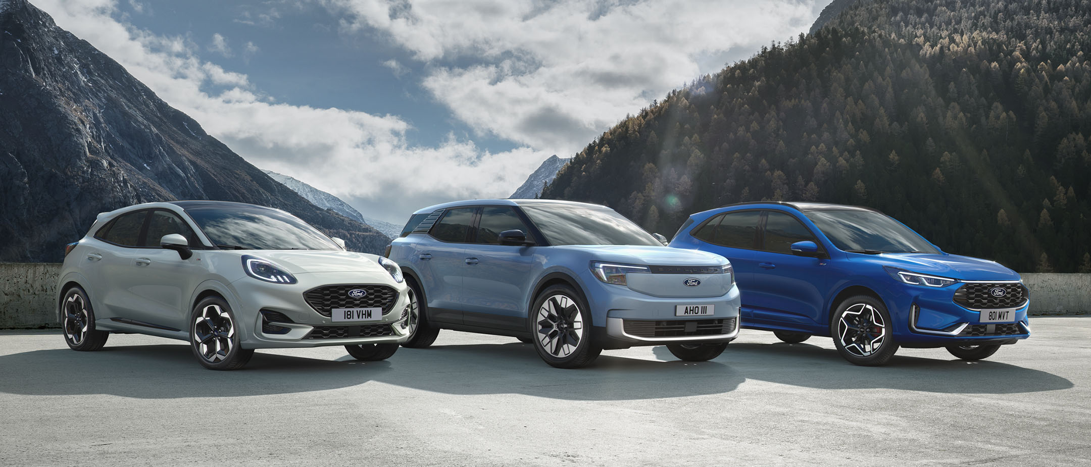 Ford Puma, Ford Explorer und Ford Kuga stehen nebeneinander auf einem Parkplatz mit Berglandschaft im Hintergrund.