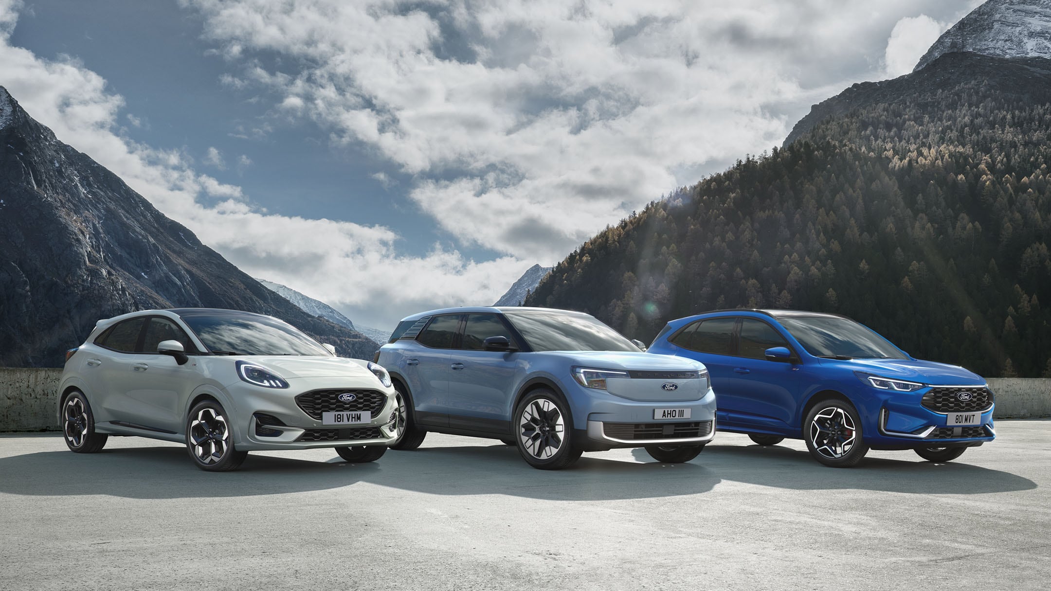 Ford Puma, Ford Explorer und Ford Kuga stehen nebeneinander auf einem Parkplatz mit Berglandschaft im Hintergrund.