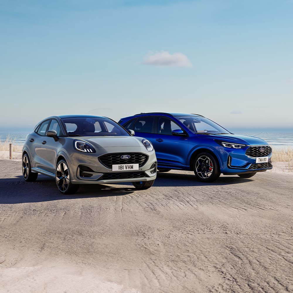 Ein grauer Ford Puma und blauer Ford Kuga parken nebeneinander auf einem Parkplatz neben dem Meer.