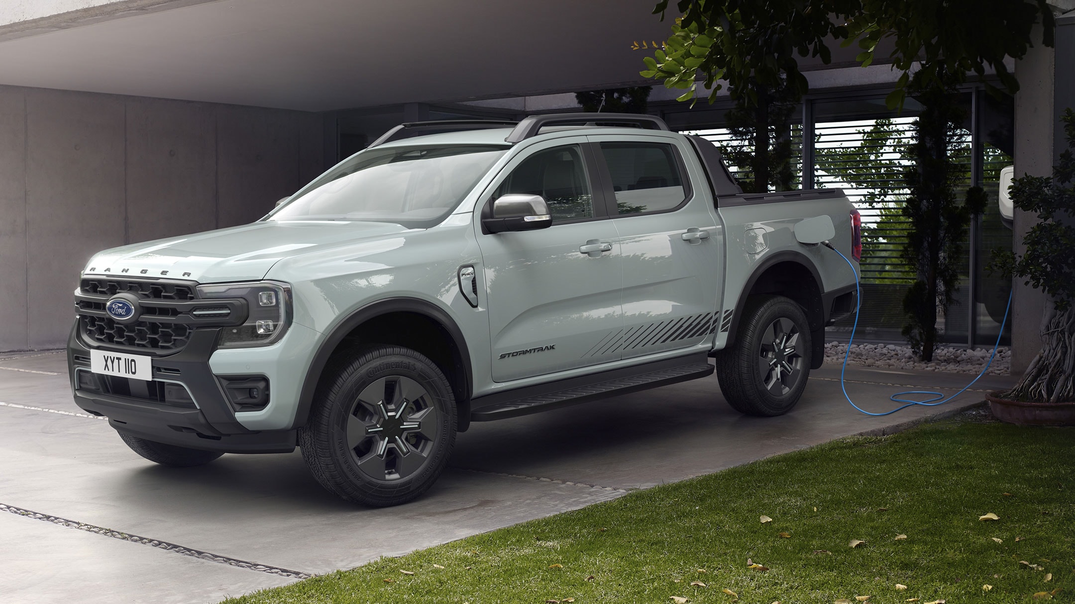 Ford Ranger Plug-in-Hybrid in Grau. Dreiviertelansicht, parkend vor einem Wohnhaus. Mann mit Hund gehen an Fahrzeug vorbei.