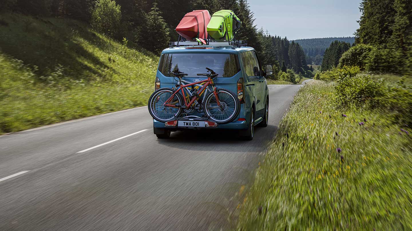 Ford Tourneo Custom in Blau. Heckansicht, fahrend auf einer Landstraße.