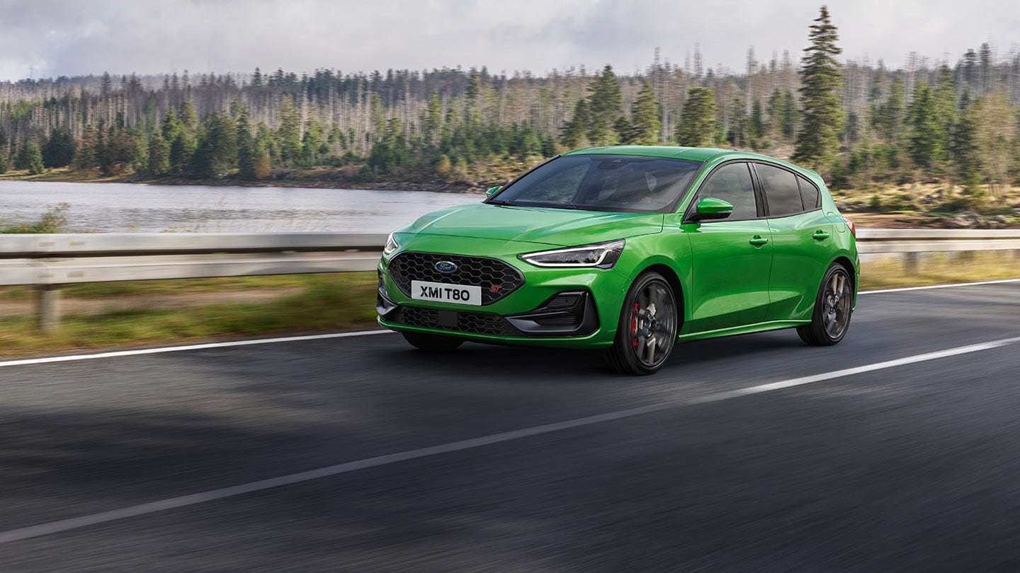 Ford Focus ST in Grün. Dreiviertelansicht, fahrend auf einer Straße. Bäume im Hintergrund.
