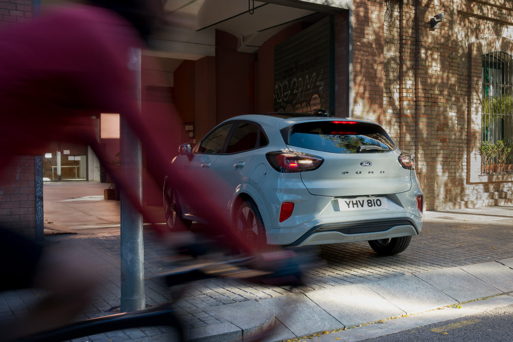 Ford Puma in Grau. Heckansicht, parkt vor einem Gebäude. Fahrradfahrer im Vordergrund.