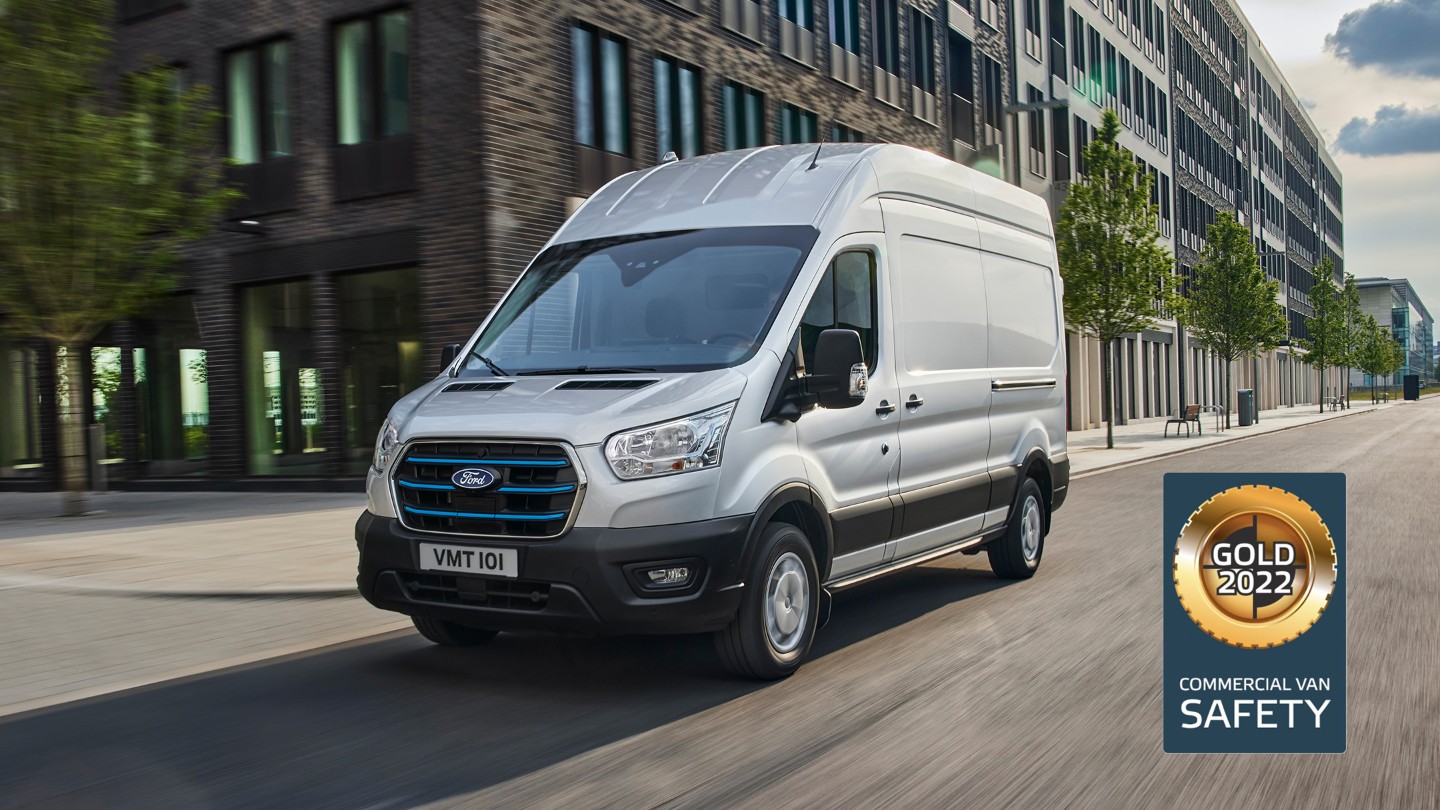 Ford E-Transit Kastenwagen in Silber. Dreiviertelansicht, fährt entlang Hochhäusern und NCAP Gold-Logo für Kategorie „Sicherheit“