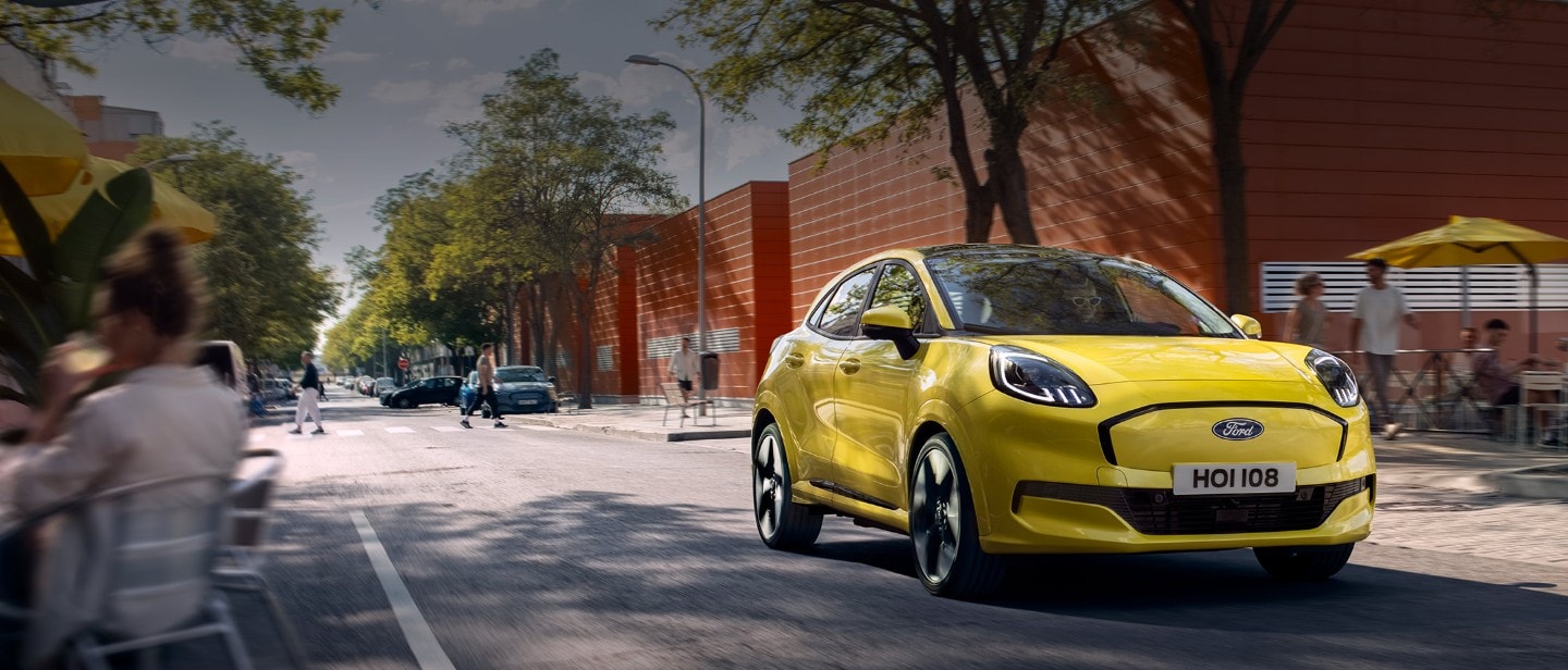 Ford Puma Gen-E in Gelb. Dreiviertelansicht, fährt auf einer Straße mit Bäumen und Personen im Hintergrund.