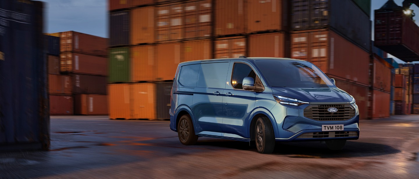 Ford Transit Custom Plug-in-Hybrid in Blau. Dreiviertelansicht, fährt vor Containern.